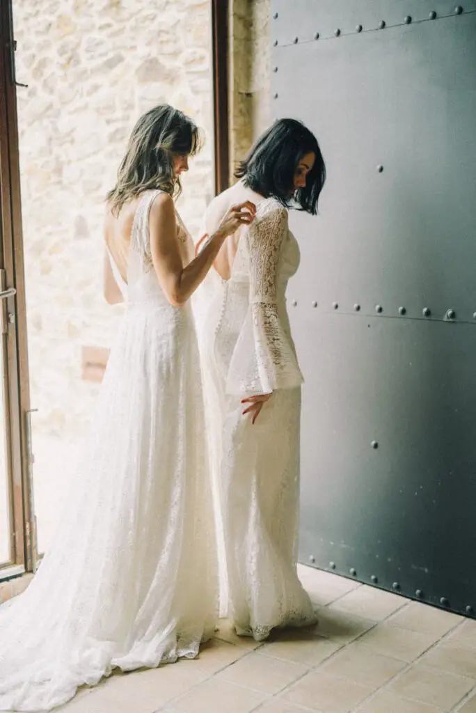 Two brides sharing a tender moment in elegant white dresses by a doorway.